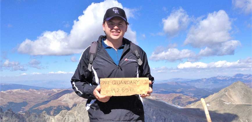 Sean A. Wirth standing on the top of Mt. Quandary holding a piece of wood with writing on it that reads, "Mt Quandary 14,265 ft.